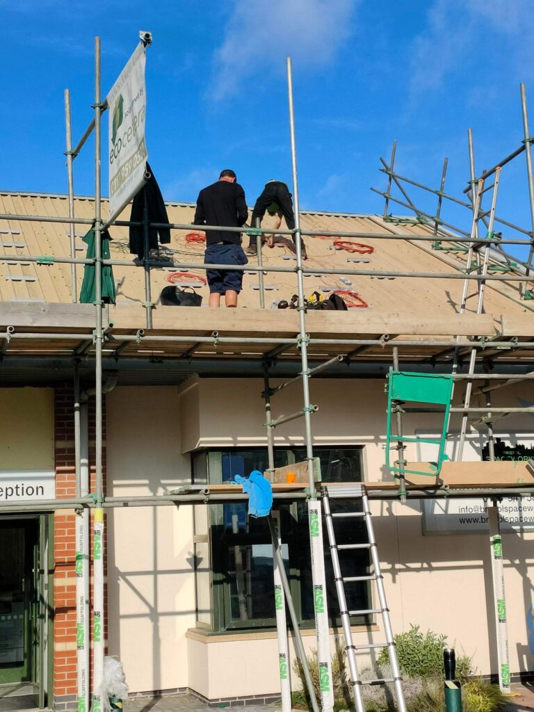 A close up of the outside of the reception building, there is scaffolding up and two people are working on installing solar panels to the roof.