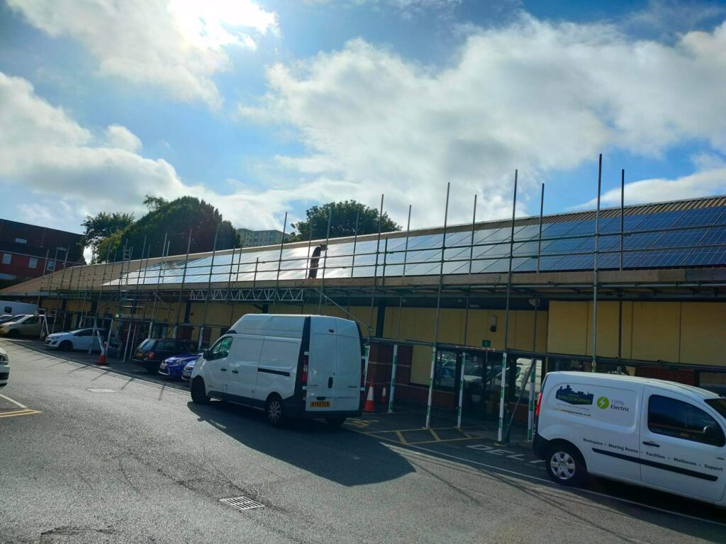 A long, one storey building with scaffolding across it. cars and vans are parked in front of the building and there is a person on top of the scaffolding doing maintenance work on the solar panels.