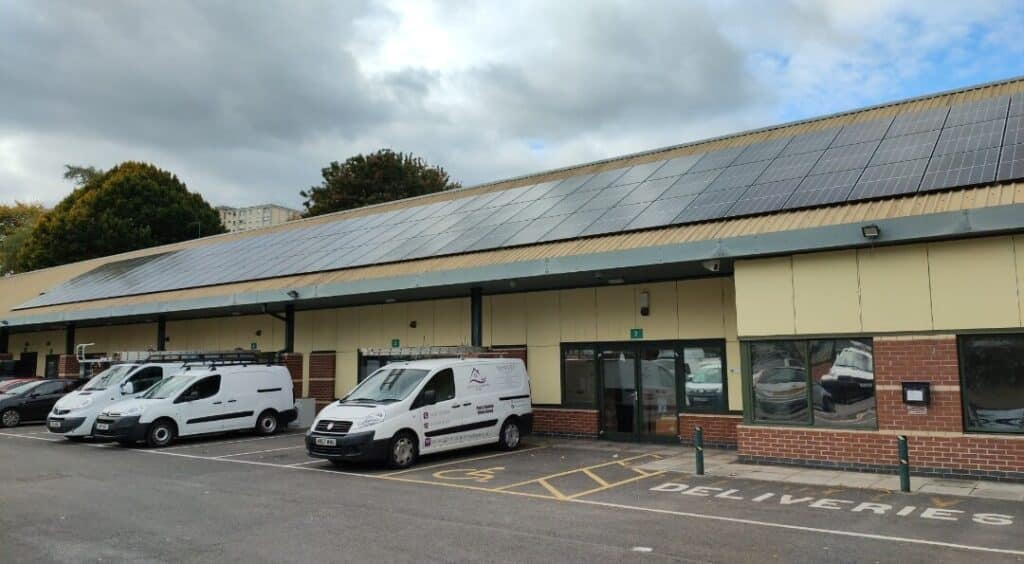 A long, one storey building with a solar panel array on top of it. There are vans parked in front of the building.