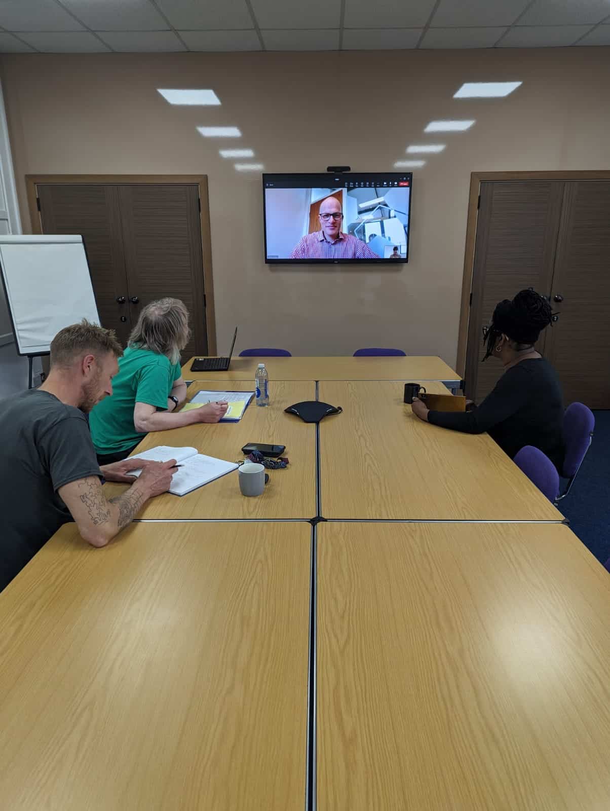 A conference room with three people seated around a table and looking towards a tv screen with a person speaking using video conferencing.