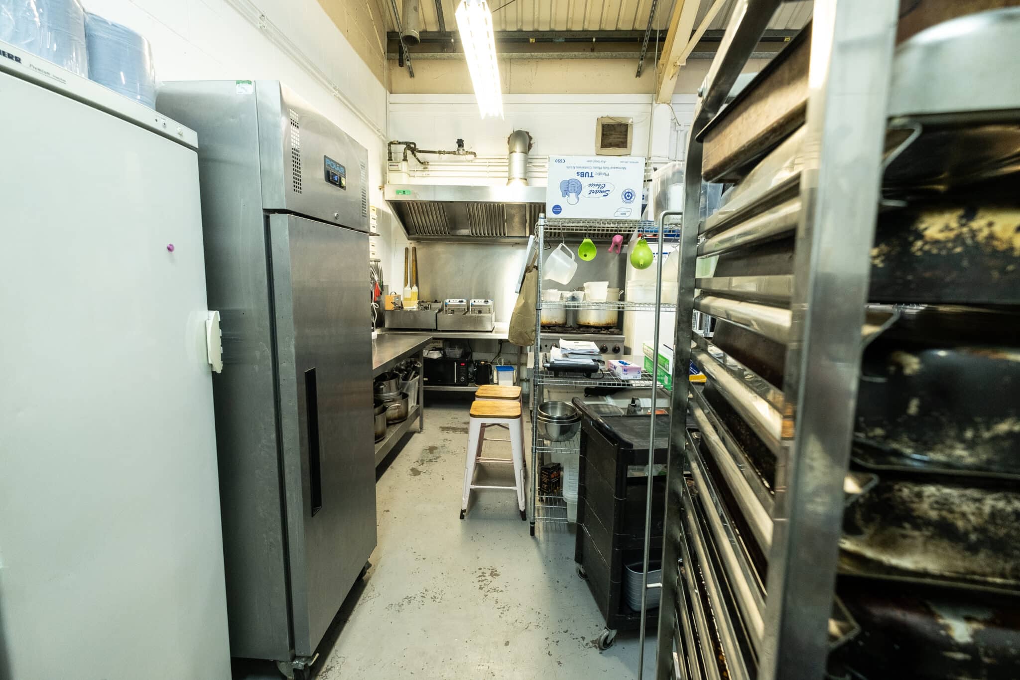 Industrial kitchen filled with metal surfaces, fridges and racks of baking trays.